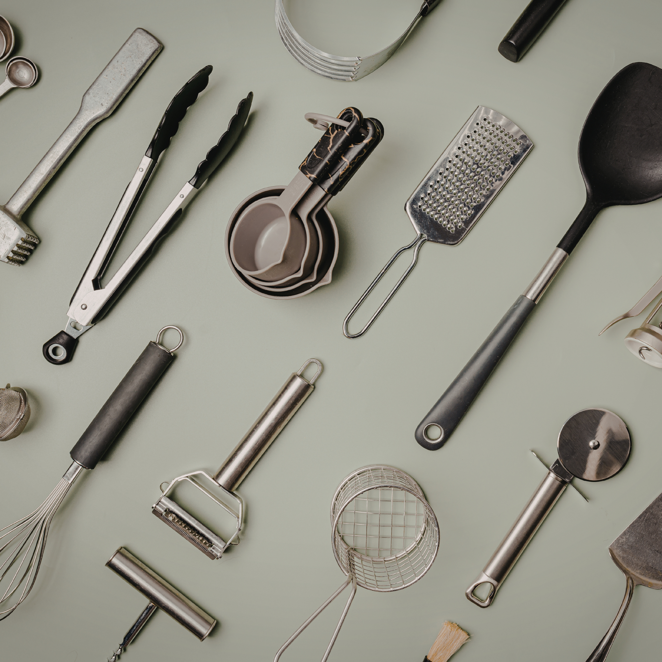 Flat lay of stainless steel kitchen tools and gadgets including tongs, ladle, grater, peeler, pizza cutter and measuring spoons on a light grey background